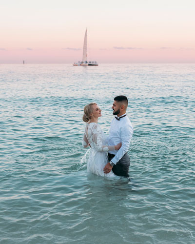 photo of a bride and groom standing in the ocean with a sailboat in the background