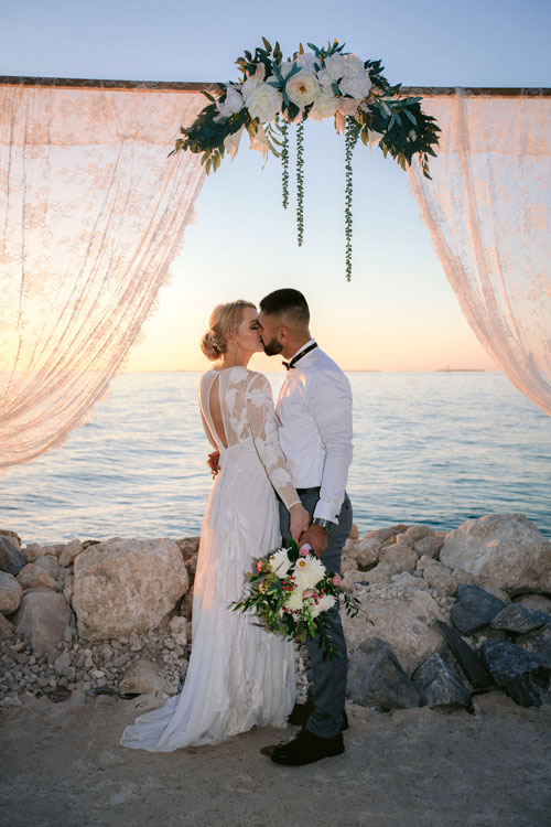 photo of a couple under a wedding arch kissing with the ocean in the background.
